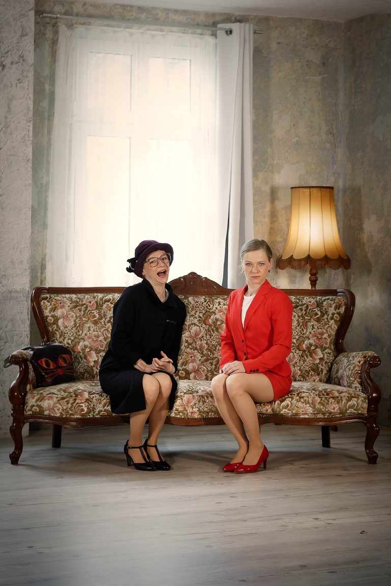 Frau Oma und Annemarie Schmidt sitzen gemeinsam auf einem schönen alten Sofa und schauen in die Kamera. Im HIntergrund eine alte Stehlampe